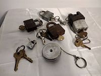 Six vintage locks and accompanying keys on a white surface, showing various designs and sizes including a round tape measure