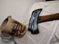 Side view of ceramic pitcher and axe head showing the painted rustic cabin and landscape on the pitcher, snowy winter scene on axe head.
