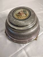 Side view of the vintage round metal powder box showing textured silver finish and lid with painted inset of woman and monkey