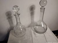Both vintage glass decanters side by side on a white cloth showing difference in design, one etched and one plain glass.