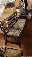 Angle view of metal frame bench with wooden slat back and patterned cushion seat, showing armrest and decorative side scrollwork.