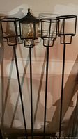 Four tall black metal garden stake lantern holders, one with a clear glass oil reservoir having a wick inside, others are empty cage holders.