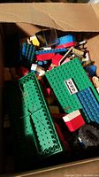 Assorted Lego blocks in a cardboard box showing different colored bricks, large green baseplates, black wheels, and transparent plastic display pieces.