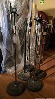 Photo showing five microphone stands grouped together on a wooden floor. The bases are round and heavy, poles are adjustable with locking mechanisms visible.
