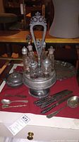 Full view of vintage silverplate cruet set with six glass bottles, covered serving bowl, serving platter, and three knives and three spoons arranged on red cloth.