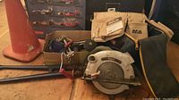 Photo showing orange safety cone, Concept circular saw, bolt cutter with red handles, Rock River tool belt, and race car poster in background.