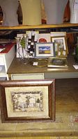 View of multiple framed artworks on table and floor including a framed sepia architectural print, smaller framed paintings, and a metal candle holder with glass votive cups in front