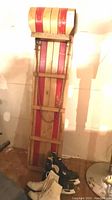 Vintage wooden toboggan standing upright with two pairs of ice skates at the bottom, showing general condition and size comparison.