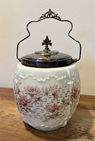 Front view of the antique biscuit jar showing white opaque glass with floral design and the silver-plated handle and lid with ornate finial.