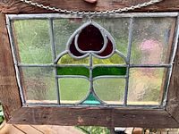 Close-up view of the stained glass window showing the red floral or heart-shaped design framed by textured clear glass and green stained glass sections. Wooden frame is visible with metal chain attached.
