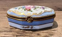Top/front angled view of antique Limoges trinket box highlighting floral hand-painted decoration, pastel blue border and gold accent highlights with metal clasp shaped like a flower.