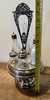 Full view of entire cruet set including the silver plated stand with ornate handle, four glass bottles inside, and a height measuring tape showing scale.