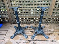 Pair of cast iron table bases on wooden deck against lattice fence, showing full height and claw feet design.