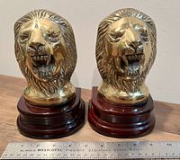 Front view of both brass lion head bookends side by side with visible measuring ruler showing height about 7 inches.