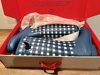 Pair of Hunter rain boots inside an open red and white box with tissue paper. Blue and white checkered pattern with solid blue accents visible.