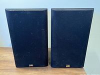 Pair of black PSB bookshelf speakers with fabric-covered fronts positioned side by side on a wood surface against a white wall.