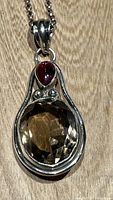 Front view of the pendant showing large oval faceted smoky quartz and smaller red teardrop cabochon stone set in sterling silver.