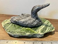 Side view of the grey stone loon sculpture sitting on a green rock base with a ruler showing length approximately 7 inches.