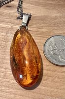 Photo showing polished Baltic amber pendant with visible natural inclusions next to Canadian quarter for size comparison on a wood surface.