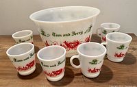 Full set with white glass punch bowl and six cups around it, showing green 'Tom and Jerry' text and red village illustration on all pieces.