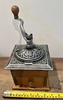 Front and top view of the vintage coffee grinder with wooden base, metal top etched with floral design, and crank handle.