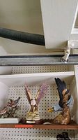 Three bird figurines displayed on a shelf with pegboard wall background. Includes eagle from The Mitella Collection with hollow head and another eagle figurine with wings spread and perched on a branch base.