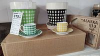 Photo shows two plastic pots with matching saucers in green and black plaid/checker design and porcelain demitasse cups/saucers with green and yellow plaid pattern, plus boxed Alaska Ulu knife.