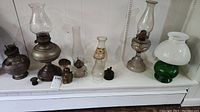 Several vintage oil lamps and hurricane lamps displayed on a white shelf with varying sizes and materials including glass chimneys, metal bases, and a white and green hurricane lamp.