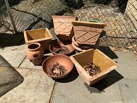 View of nine garden pots outdoors on stone patio, showing various shapes including square and round. Includes terracotta, plastic, and resin materials with leaves and dirt inside.