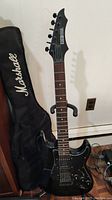 Full view of black Rocket Special electric guitar with Marshall branded soft case beside it on the floor, guitar on stand.