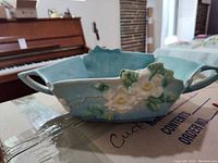 Side view of the Blue Roseville ceramic serving dish with white and yellow flowers and green leaves on one side. Two handles visible. Cracks present.