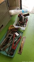 Photo showing various hand saws, a large red bolt cutter, metal clamps, small hand tools in a plastic container, and a roll of white plastic sheeting.