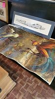 Photo of the two pieces of wall art together, showing the colorful cock fight painting in front and the black and white Chapel Street print behind it.