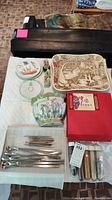 Top-down view of lot items on white quilted surface including ceramic wall pocket, plates, carved tray, utensils, figurines, and boxes