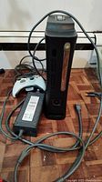 Xbox 360 console upright with controller, power supply, and AV cables visible on wood floor.