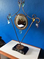 Full view of the Art Deco mirror showing circular mirror, metal frame, colored glass leaf-shaped accents hanging from scrolls, and blue glass or stone base.