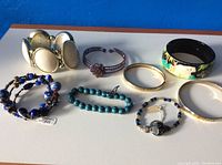 Overview photo showing all eight bracelets laid out on blue background, including painted animal bangle, glass bead bracelets, and cream and metal toned bangles.