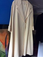 Full view of the long cream coat jacket hanging on a hanger showing fabric, lapels, and overall design.