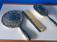 Set of vintage dresser items showing brush, mirror, and comb with detailed silver tone floral design.