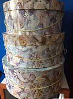 Stack of three round hat boxes with floral and butterfly patterns on a blue background. Yellow cords wrapped around each box.