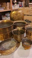 Photo showing multiple brass planters and the tall brass vase in the background, arranged on a surface with boxes behind.