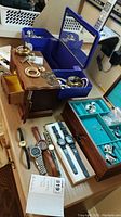 Overview image showing wood jewellery boxes with blue lining and a mirror, various costume jewellery and watches arranged on a table.