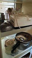 Top shelf view showing white Cuisinart food processor with attached power cord, stainless steel steamer basket, and parts of juicer and pressure cooker.