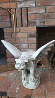 Front view of the concrete gargoyle plant holder showing its detailed face, wings, and posture on a brick fireplace hearth.