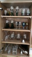 Multiple shelves shown containing clear etched stemmed wine glasses with green stems, colored goblets, printed tumblers, assorted clear stemless glasses, and a silver metal cocktail shaker.