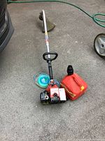 Full view of the Homelite Bandit gas trimmer lying on the ground alongside a blue trimmer line spool and a red gas can.