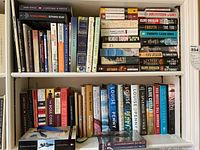 Two shelves filled with hardcover and trade edition paperback novels featuring authors Clive Cussler, Louise Penny, Robert Ludlum, and others.