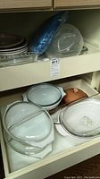 Shelf with stacked glass pie plates, glass lasagna pans, Corning Ware casseroles with lids, clay roaster underneath.
