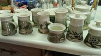 Eleven pieces including six handmade pottery mugs with three different landscape designs and a teapot on counter.