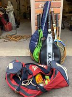 Full view of collection with racquets in covers, carry bag containing balls and jump rope in front, grouped outdoors on pavement near a garage.